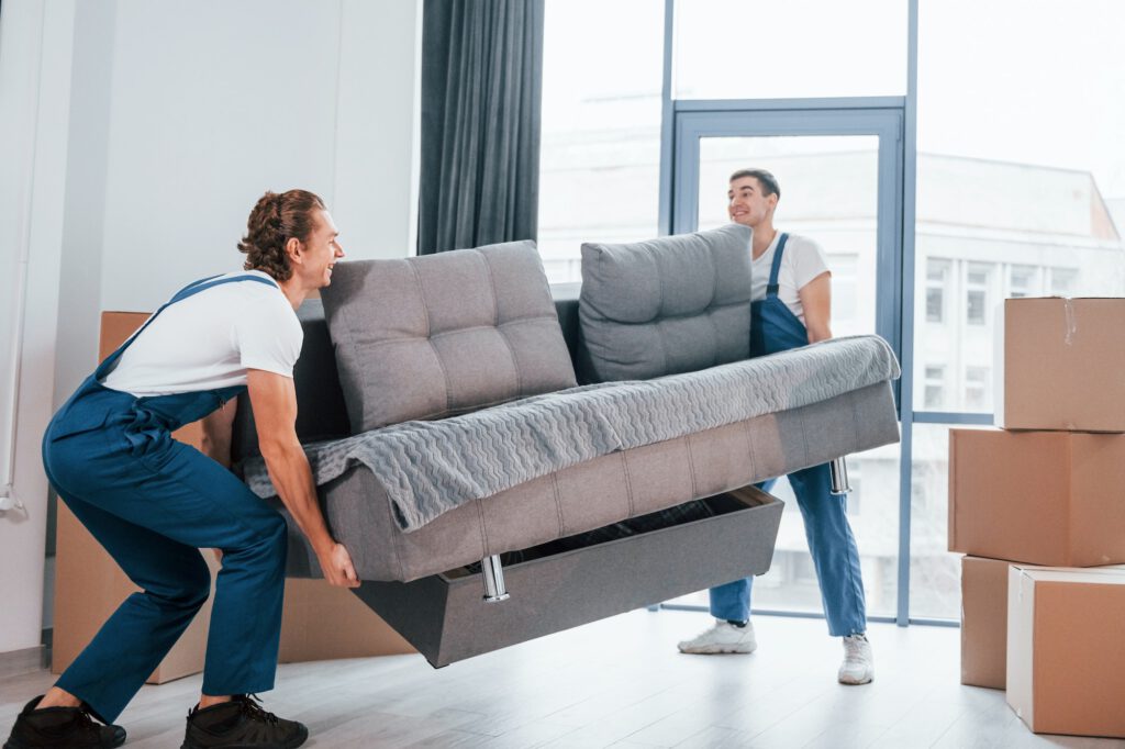 Carrying heavy sofa. Two young movers in blue uniform working indoors in the room